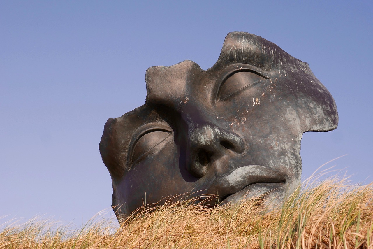 large weathered mask sculpture partially buried in sand representing distorted digital identity